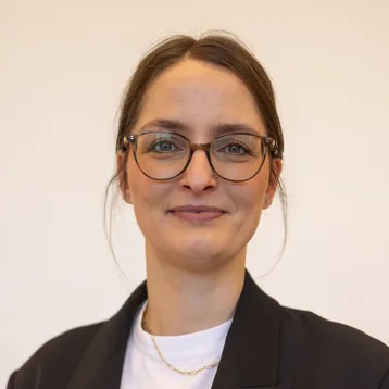 Portrait von Hanna Klein vor hellem Hintergrund, sie trägt ein Jackett und lächelt in die Kamera 