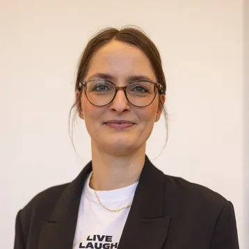 Portrait von Hanna Klein vor hellem Hintergrund, sie trägt ein Jackett und lächelt in die Kamera 