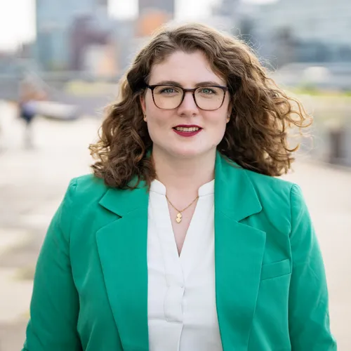 Franziska Hollstein steht vor einer undeutlichen Stadt-Kulisse und schaut selbstbewusst in die Kamera 